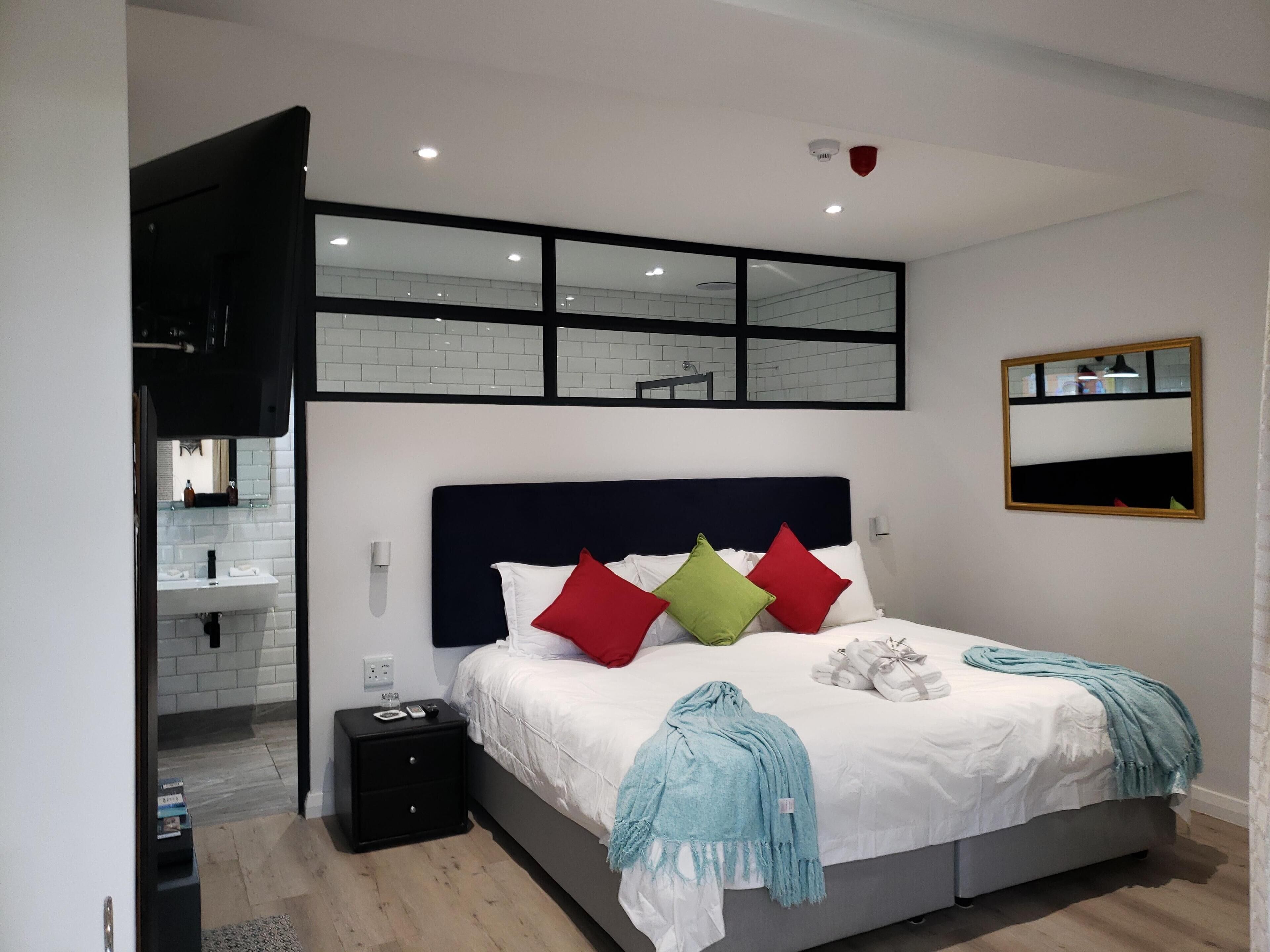 Quarto moderno com uma cama grande, mesa de cabeceira, espelho e banheiro visível através de um painel de vidro.