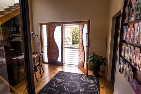 A hallway with a wooden floor, a desk with chairs, a bookshelf, and a door leading to a garden. - Point B Guest House (Cape Town)