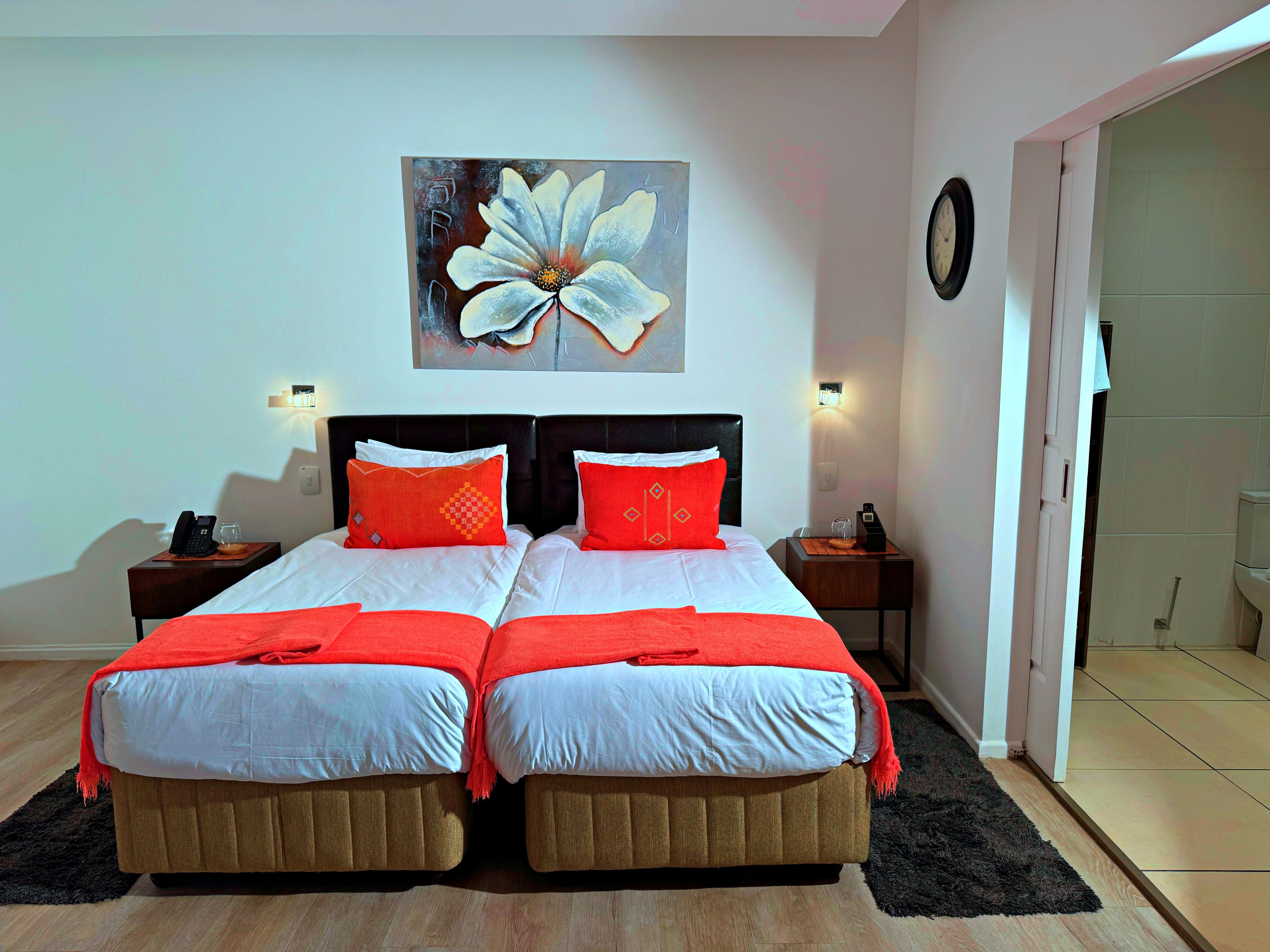 Un lit double avec du linge de lit blanc et des touches de rouge, un tableau représentant une fleur accroché au mur et une horloge également fixée au mur.