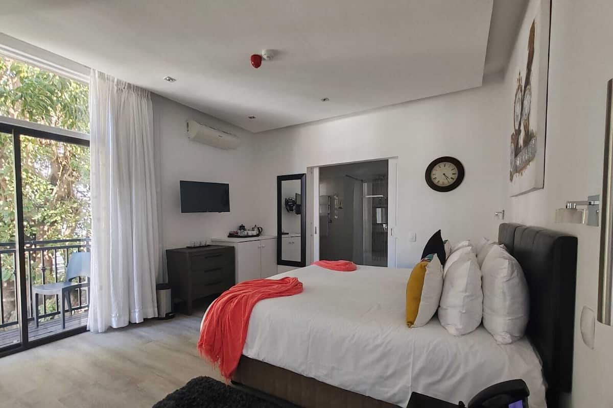 A modern bedroom with a large bed, a TV, a mirror, and a balcony with a view of trees.