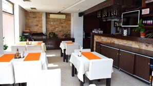A restaurant with tables covered in white tablecloths and orange placemats, a stone wall, and a bar area with a television and bar stools. - Hotel Elitsa (Bansko)