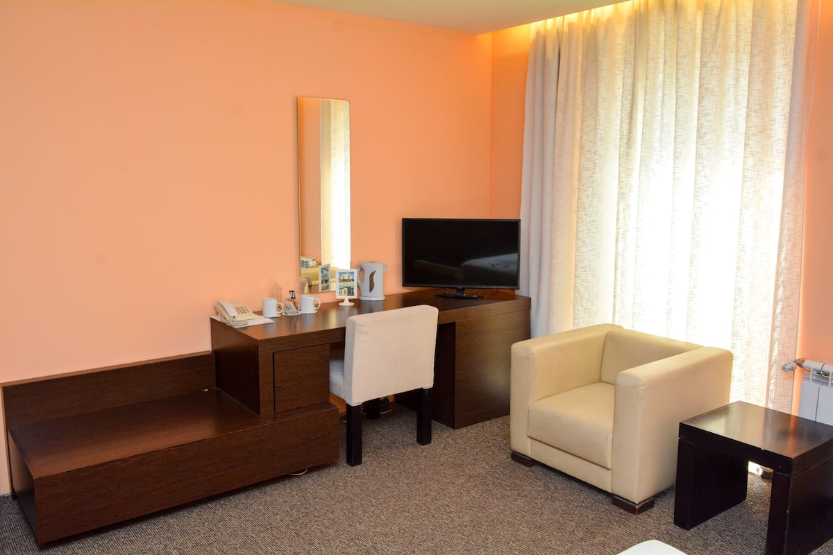 A hotel room with a desk, a chair, a sofa, a TV, and a large window with curtains.