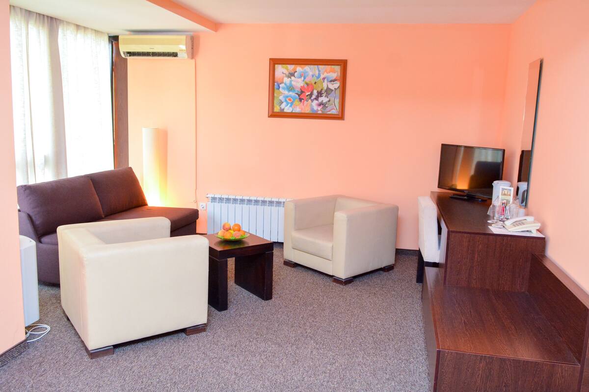 A hotel room with a brown sofa, a white armchair, a small table with fruit, a wooden desk, a television, and a painting on the wall.