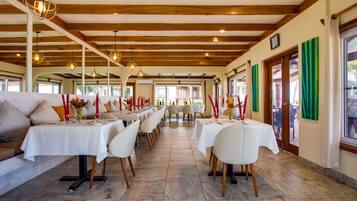 A restaurant with long tables, white chairs, and hanging pendant lights.