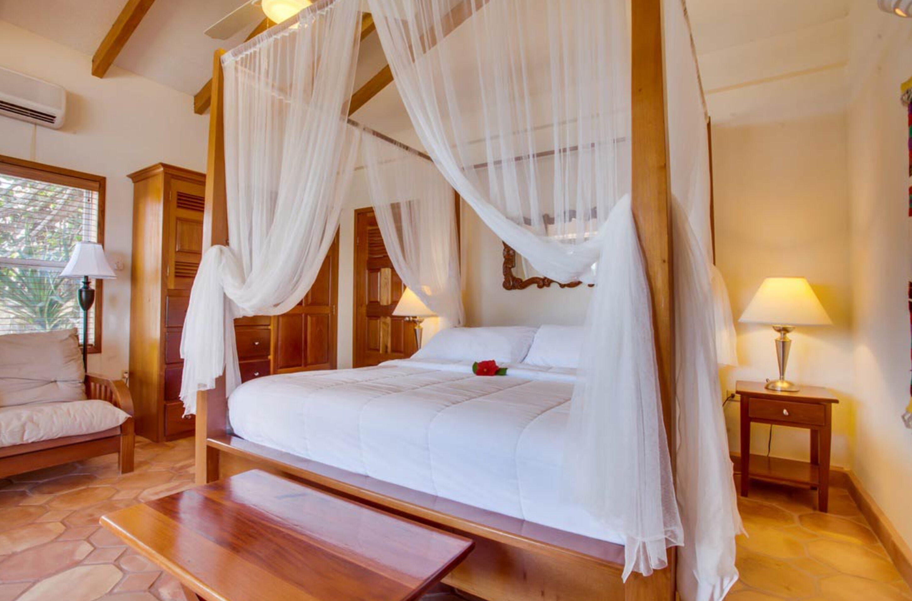 A four-poster bed with white linens and a canopy, a wooden bench, a bedside table with a lamp, and a wooden nightstand with another lamp.