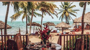 A wooden deck with a table and chairs overlooking a beach with palm trees and thatched umbrellas. - Belizean Dreams Resort (Hopkins)