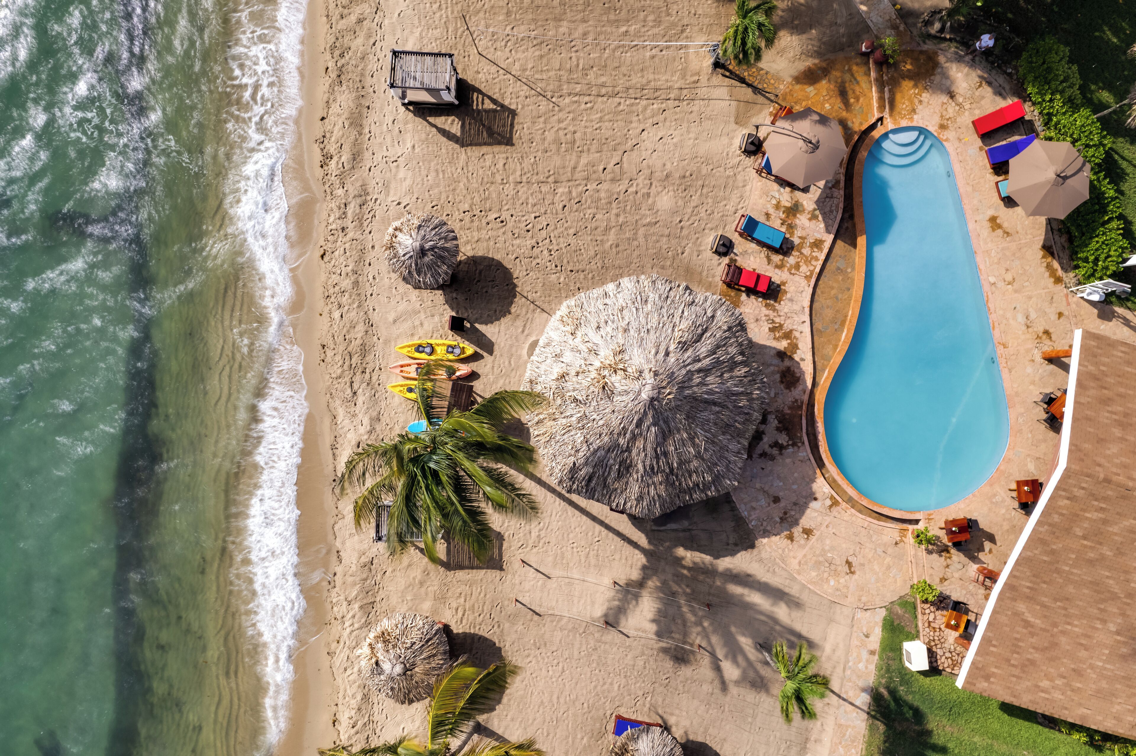 Vista aérea de una playa con piscina, sombrillas, sillas de descanso y palmeras.