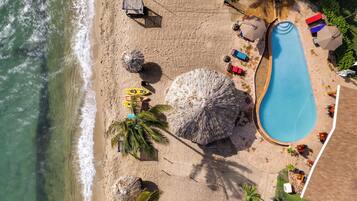 Vista aérea de una playa con piscina, sombrillas, sillas de descanso y palmeras.