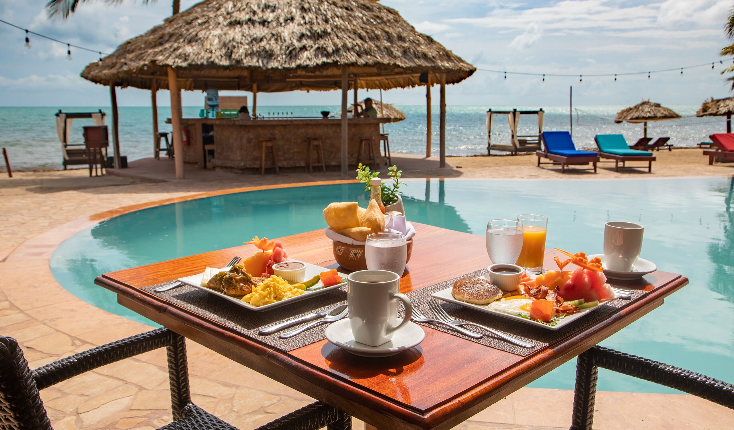 A poolside breakfast table with a variety of food and drinks, overlooking a beach with lounge chairs and a bar area.