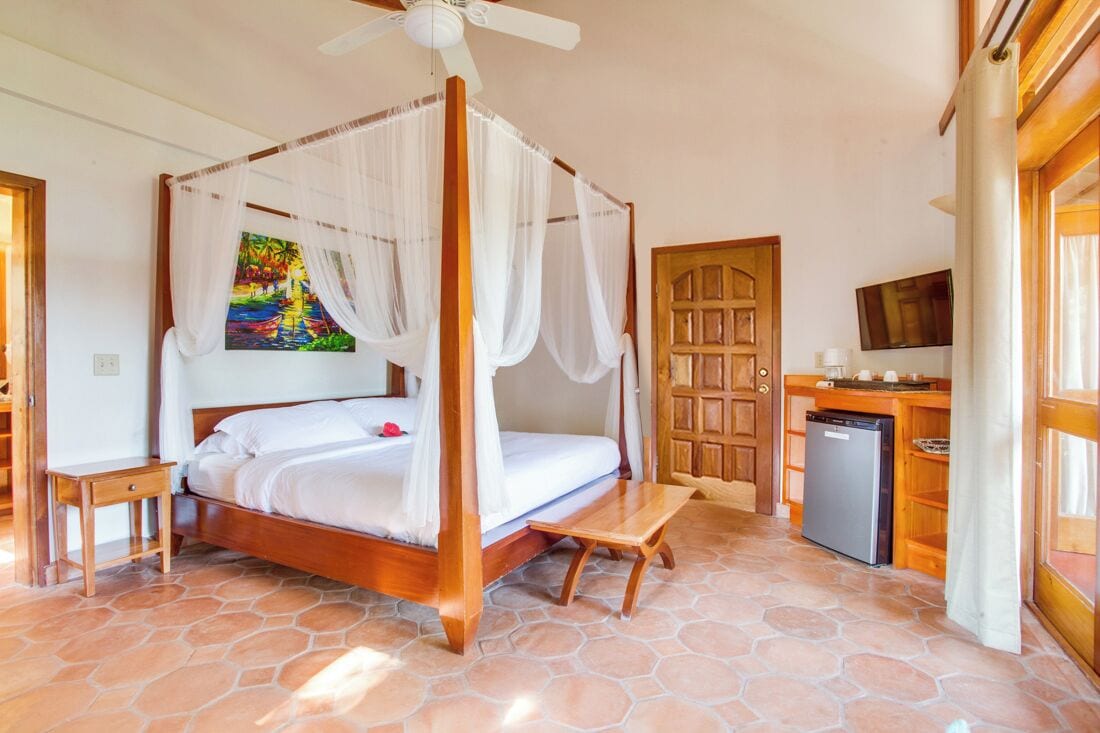 A bedroom with a four-poster bed, a television, a small kitchen area, and a stone-tiled floor.
