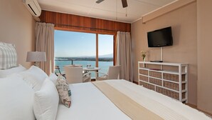 Habitación de hotel con una cama grande, un televisor montado en la pared, una pequeña mesa con sillas y vista al mar por la ventana.