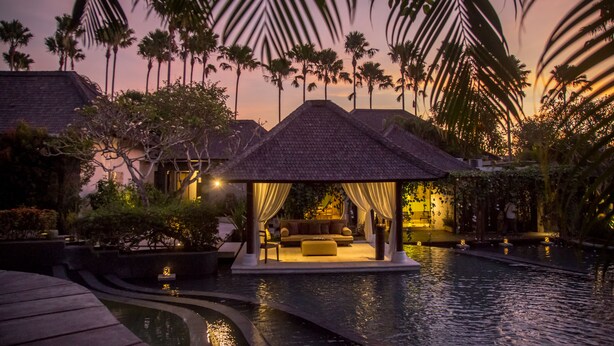 Um ambiente tranquilo ao entardecer, com piscina, área de estar coberta e palmeiras.