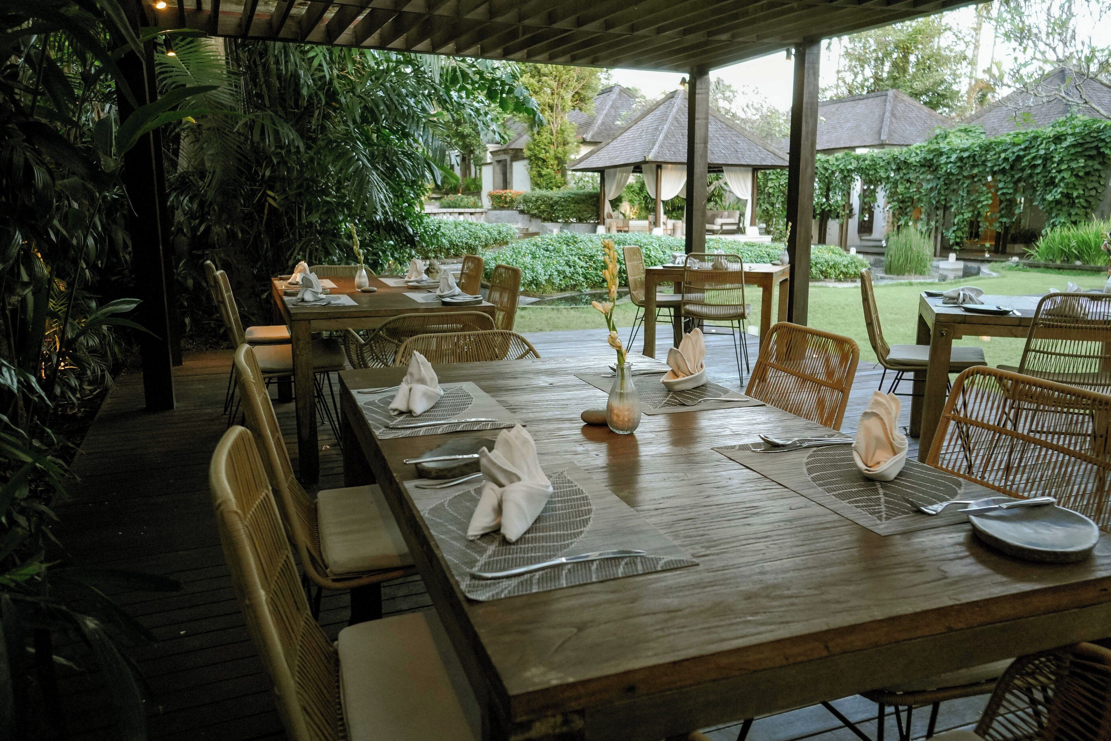 A covered outdoor dining area with wooden tables and wicker chairs, surrounded by lush greenery and a view of traditional buildings.