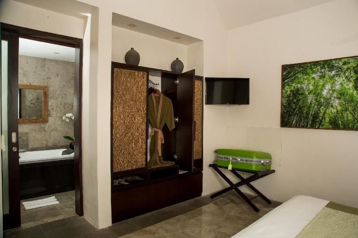 Quarto de hotel com guarda-roupa, mala verde, televisão e pintura de uma floresta.