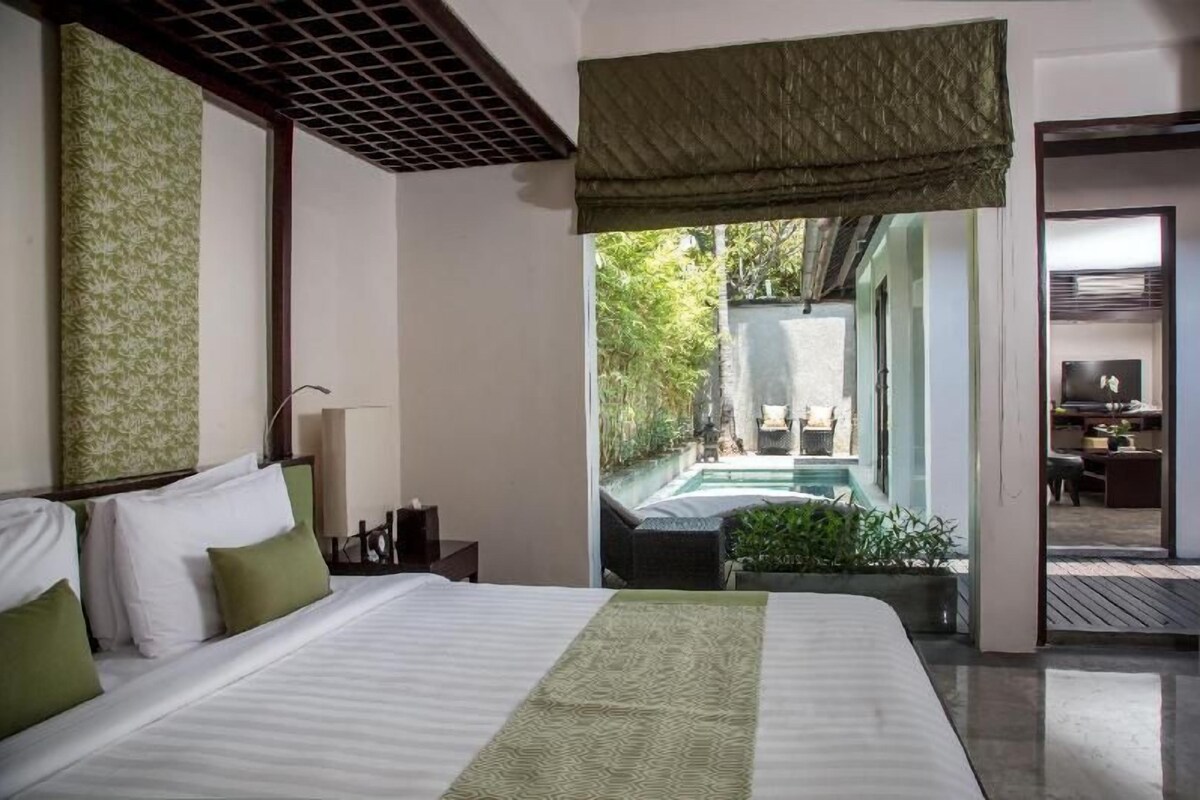 Quarto de hotel com cama, mesa de cabeceira, luminária, vista para uma piscina ao ar livre e um vislumbre de uma área de refeições.