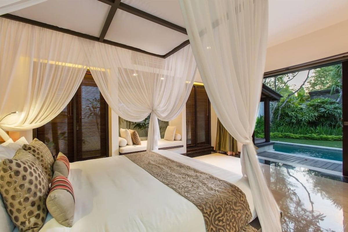 Une chambre avec un grand lit, des rideaux blancs, une vue sur une piscine et un coin salon avec des coussins.