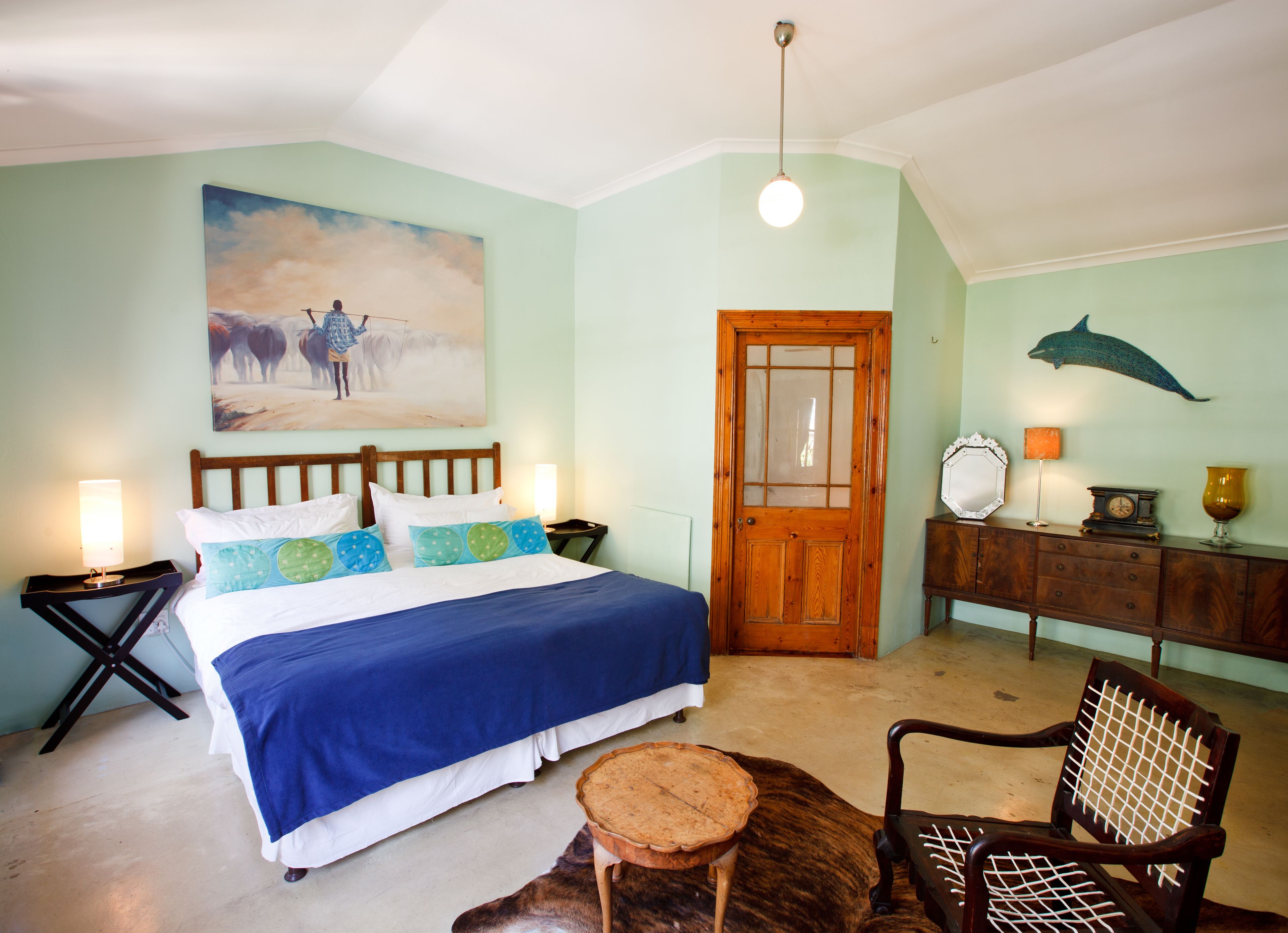 A bedroom with a bed, a wooden door, a painting of cows, a lamp, a clock, and a dolphin wall decoration.