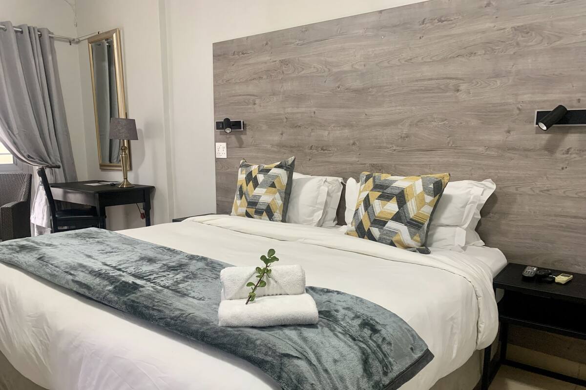 Una cama bien hecha con edredón gris y blanco, dos almohadas estampadas, una planta pequeña y una mesita de noche con lámpara y teléfono.