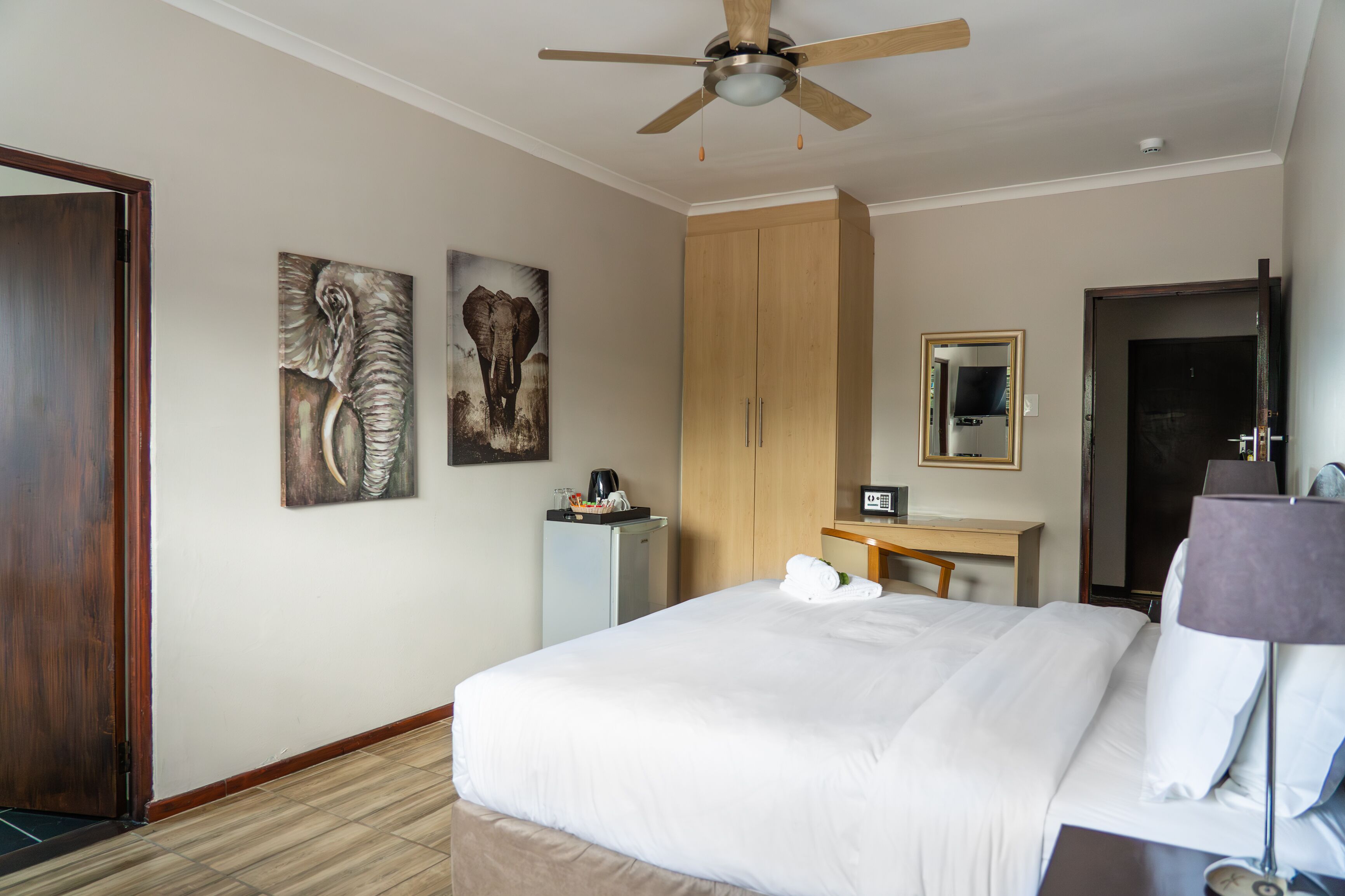 A hotel room with a bed, a desk, a wardrobe, a ceiling fan, and artwork on the walls.