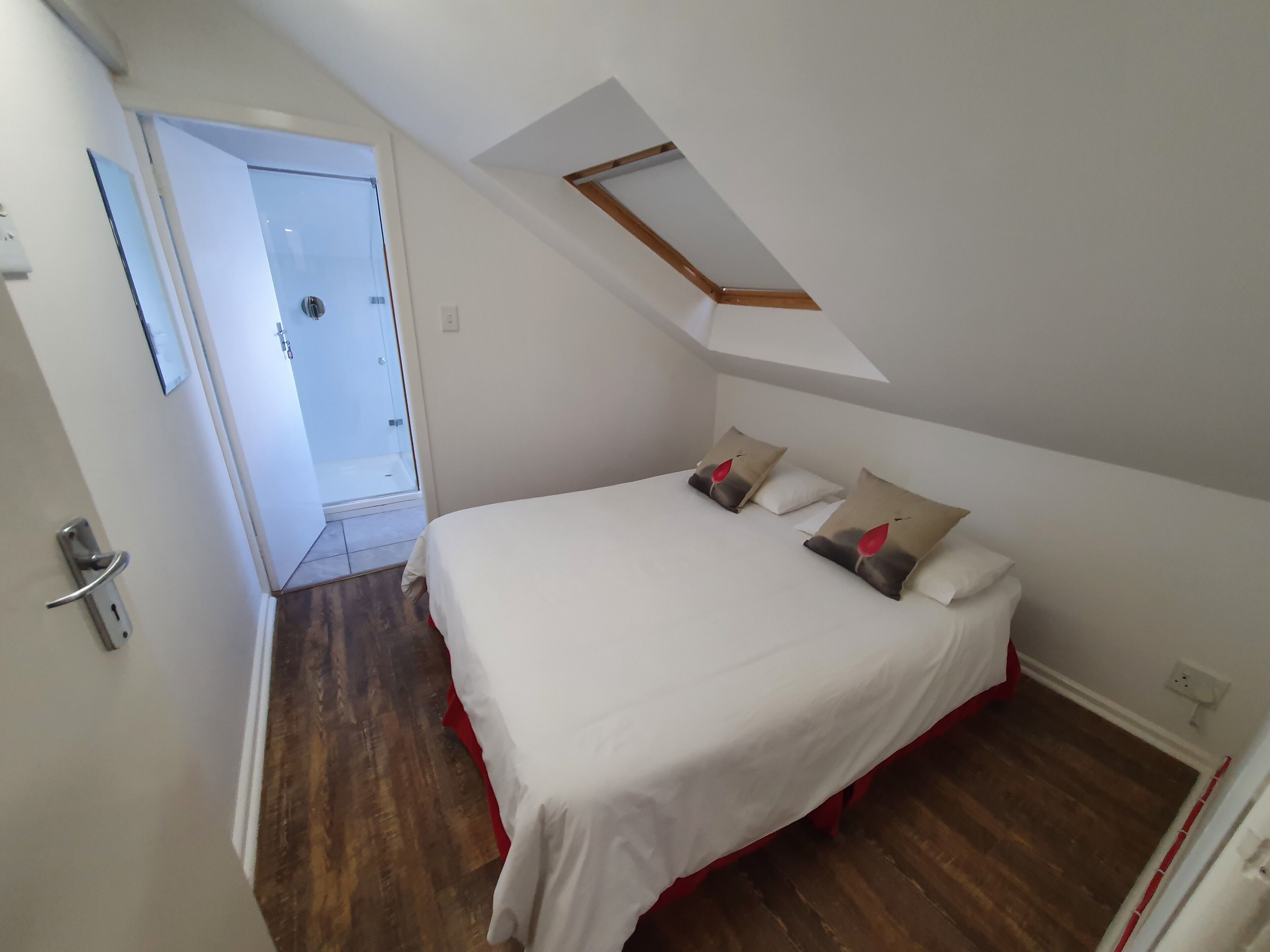 Een slaapkamer met een schuine kap, een dakraam, een bed met witte beddengoed en decoratieve kussens, en een houten vloer.