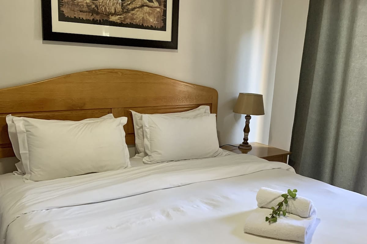 Una cama bien hecha con ropa blanca, cabecera de madera y un cuadro enmarcado encima.