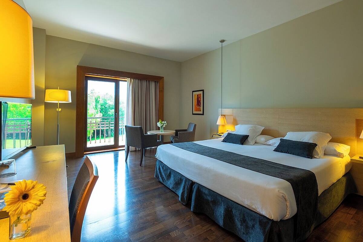 Een hotelkamer met een groot bed, een bureau met stoel, een tafeltje met bloemen en een balkon met uitzicht op groen.
