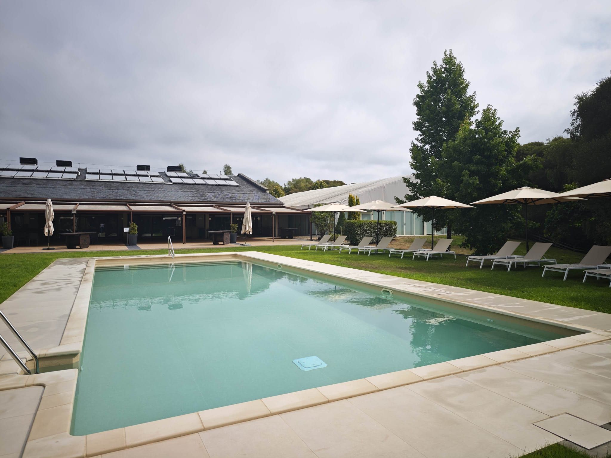 Une piscine rectangulaire avec une terrasse carrelée, entourée de chaises longues et de parasols, et un bâtiment avec un toit en pente en arrière-plan.