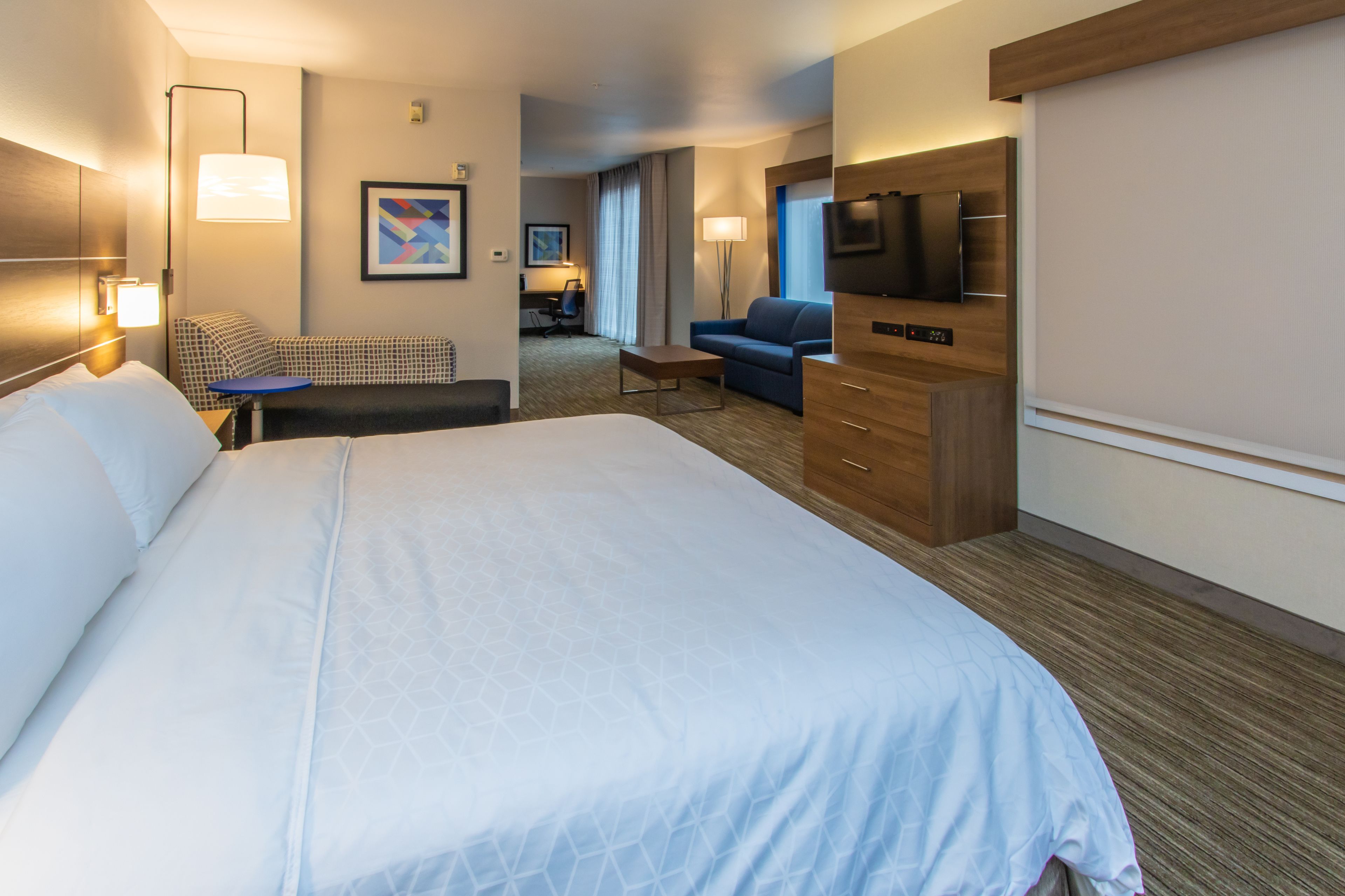 A hotel room with a large bed, a desk, a chair, a TV, and a desk lamp.