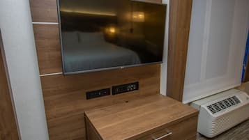 A modern hotel room with a flat-screen TV mounted on a wooden wall, a small wooden cabinet with drawers, and a portable air conditioning unit.