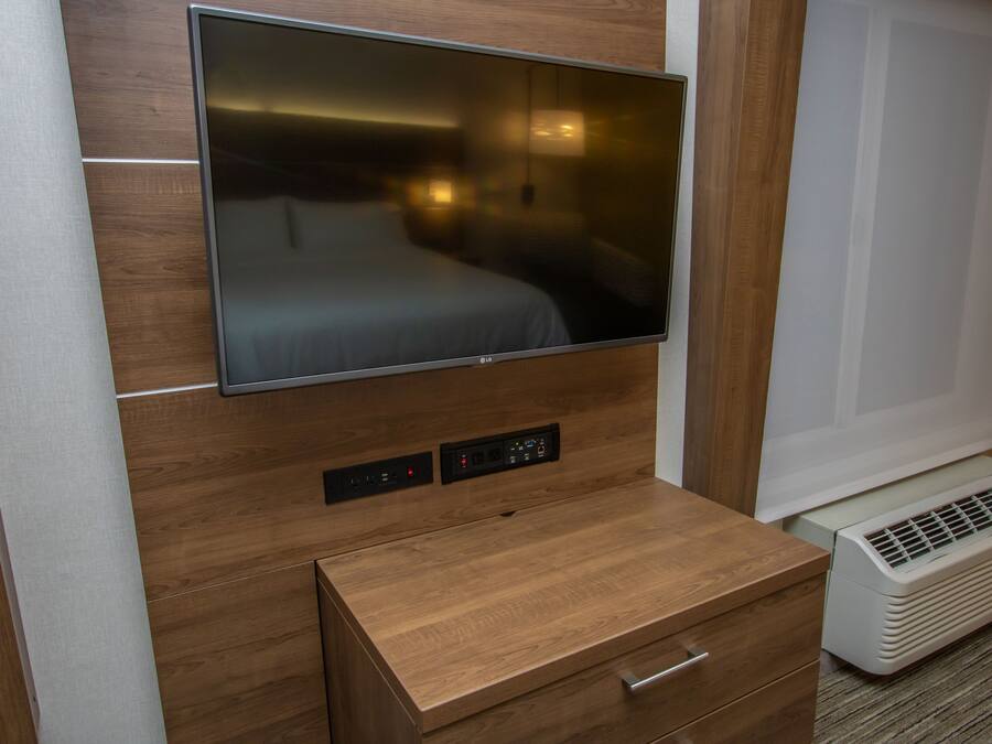 A modern hotel room with a flat-screen TV mounted on a wooden wall, a small wooden cabinet with drawers, and a portable air conditioning unit.