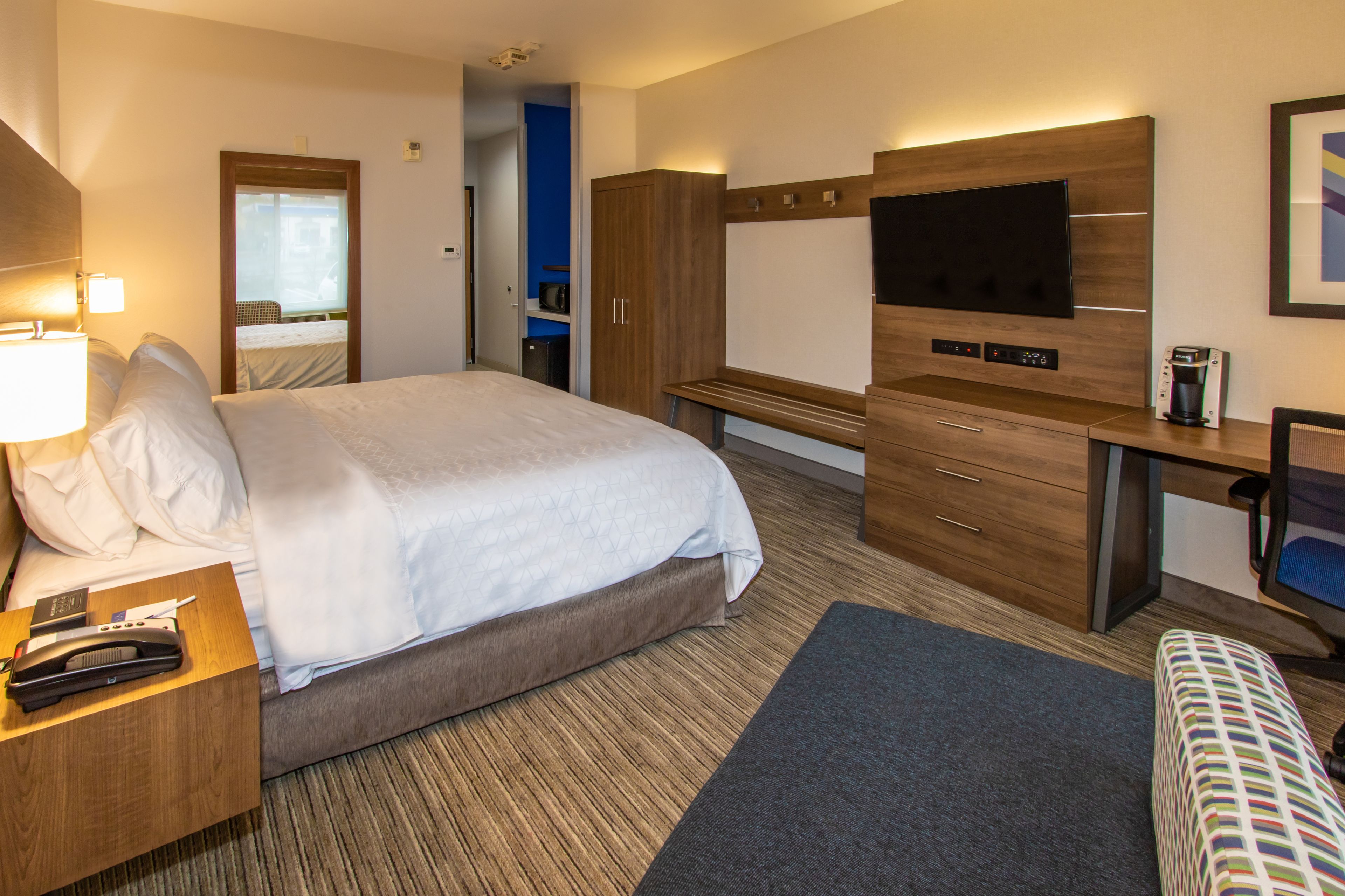 Habitación de hotel con una cama grande, mesita de noche con lámpara, escritorio con computadora, televisor y cafetera.