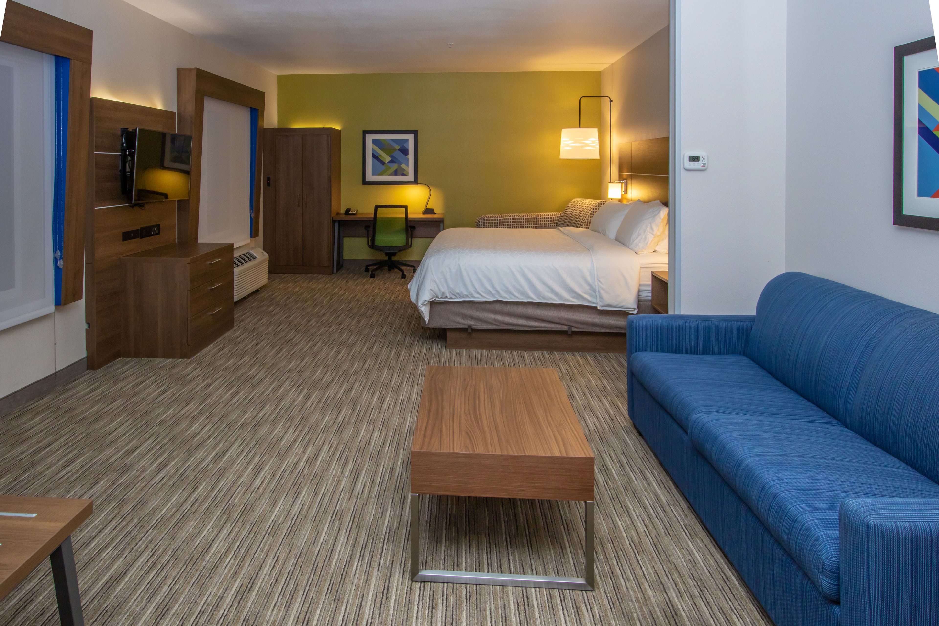 A hotel room with a bed, a blue sofa, a wooden desk, a chair, a TV, and a framed picture on the wall.