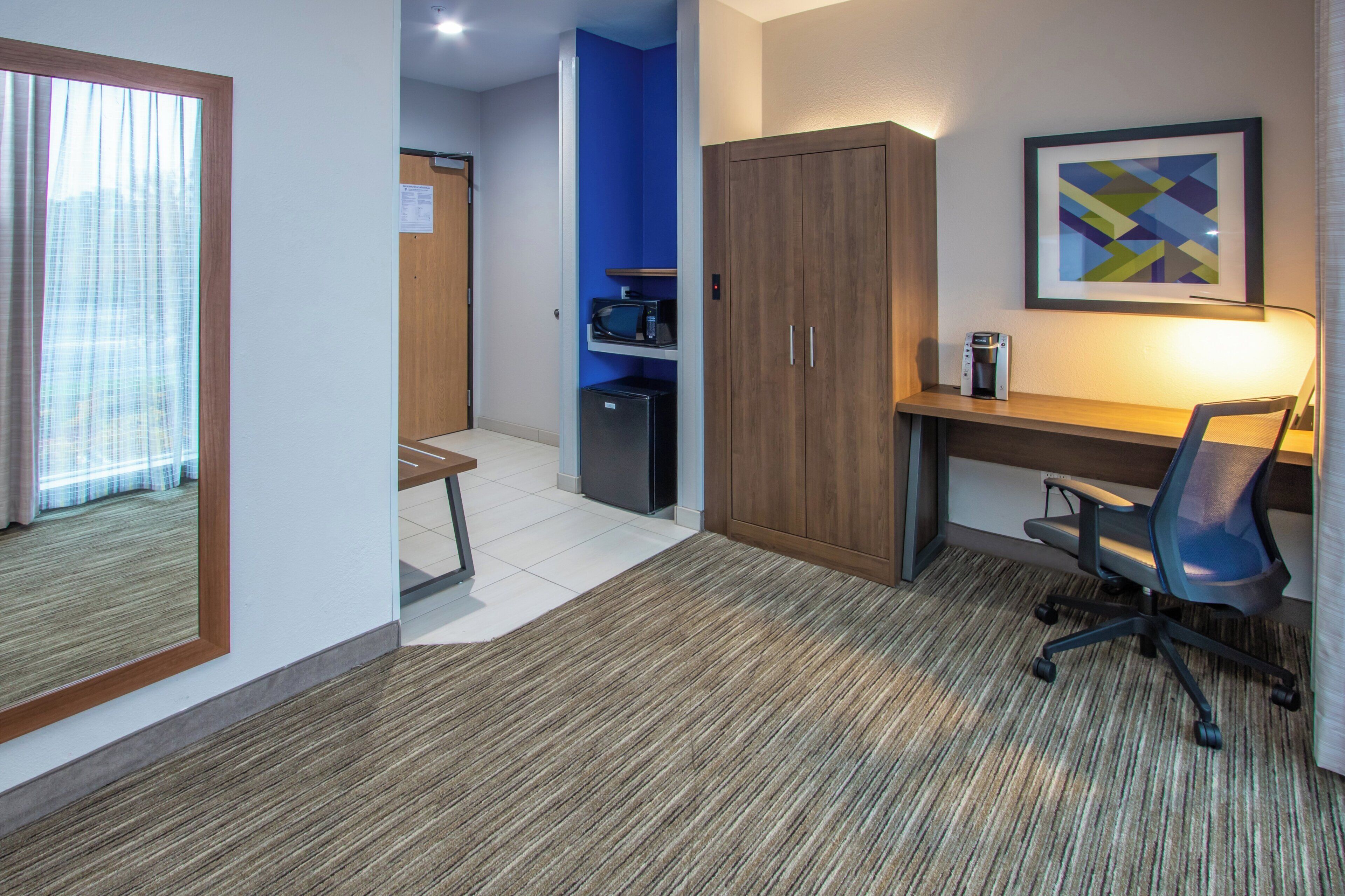 A hotel room with a desk, chair, kitchenette, and a framed picture on the wall.