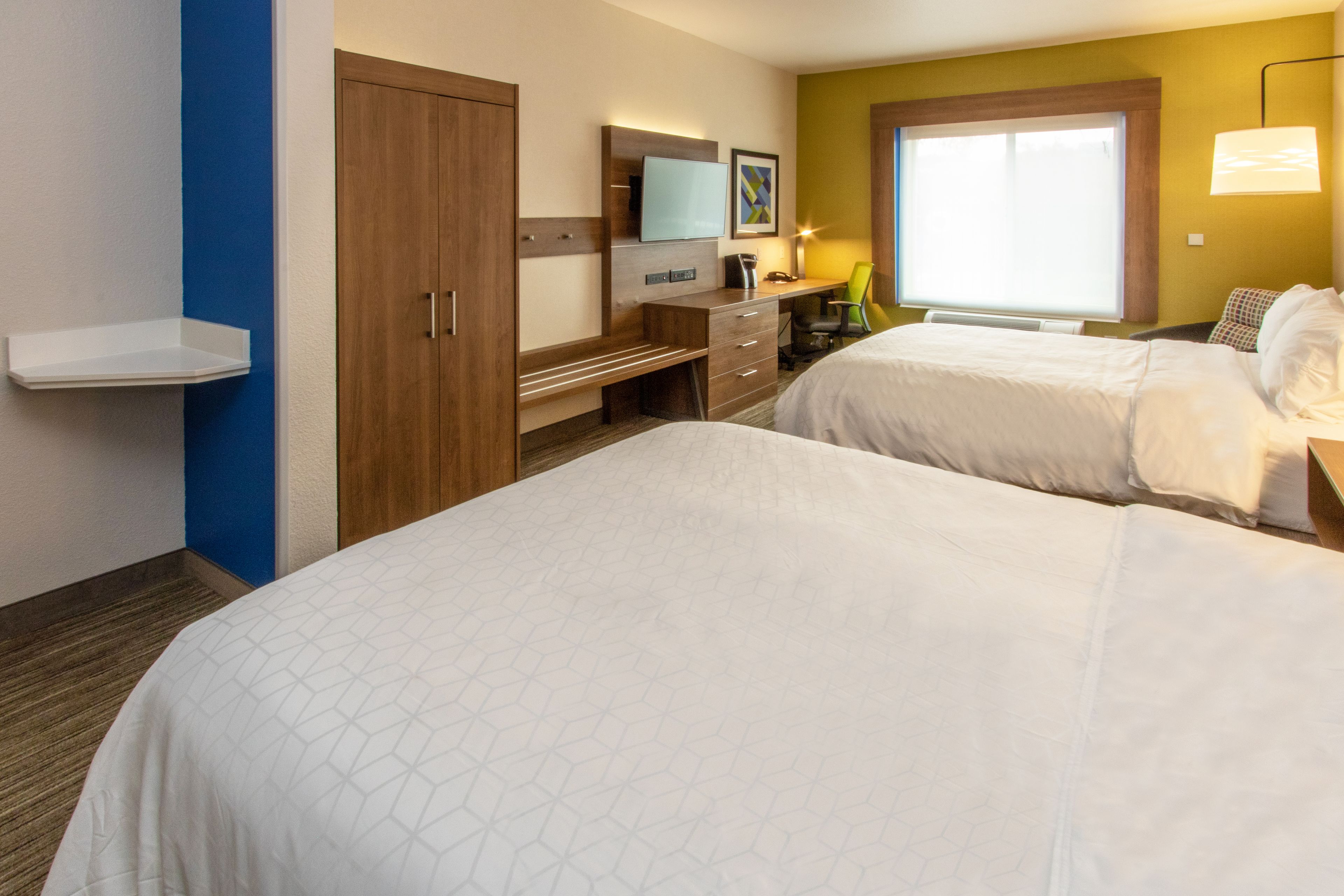 A hotel room with two beds, a desk, a chair, a TV, and a window with blinds.