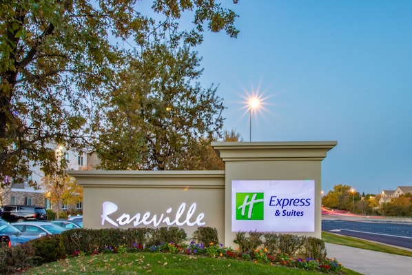 A hotel sign with 'Roseville' and 'Express & Suites' branding, surrounded by greenery and a parking area.