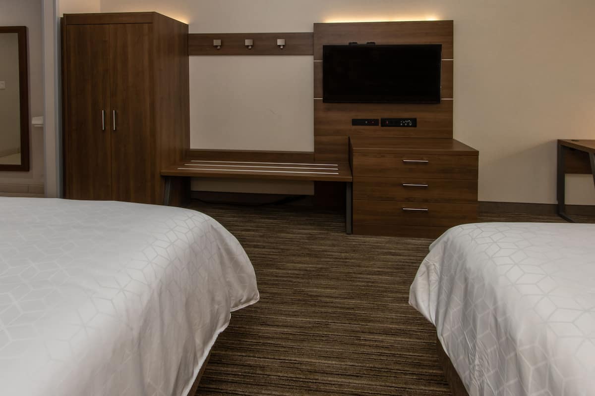 A hotel room with two beds, a wooden wardrobe, a desk with a flat-screen TV, and a carpeted floor.