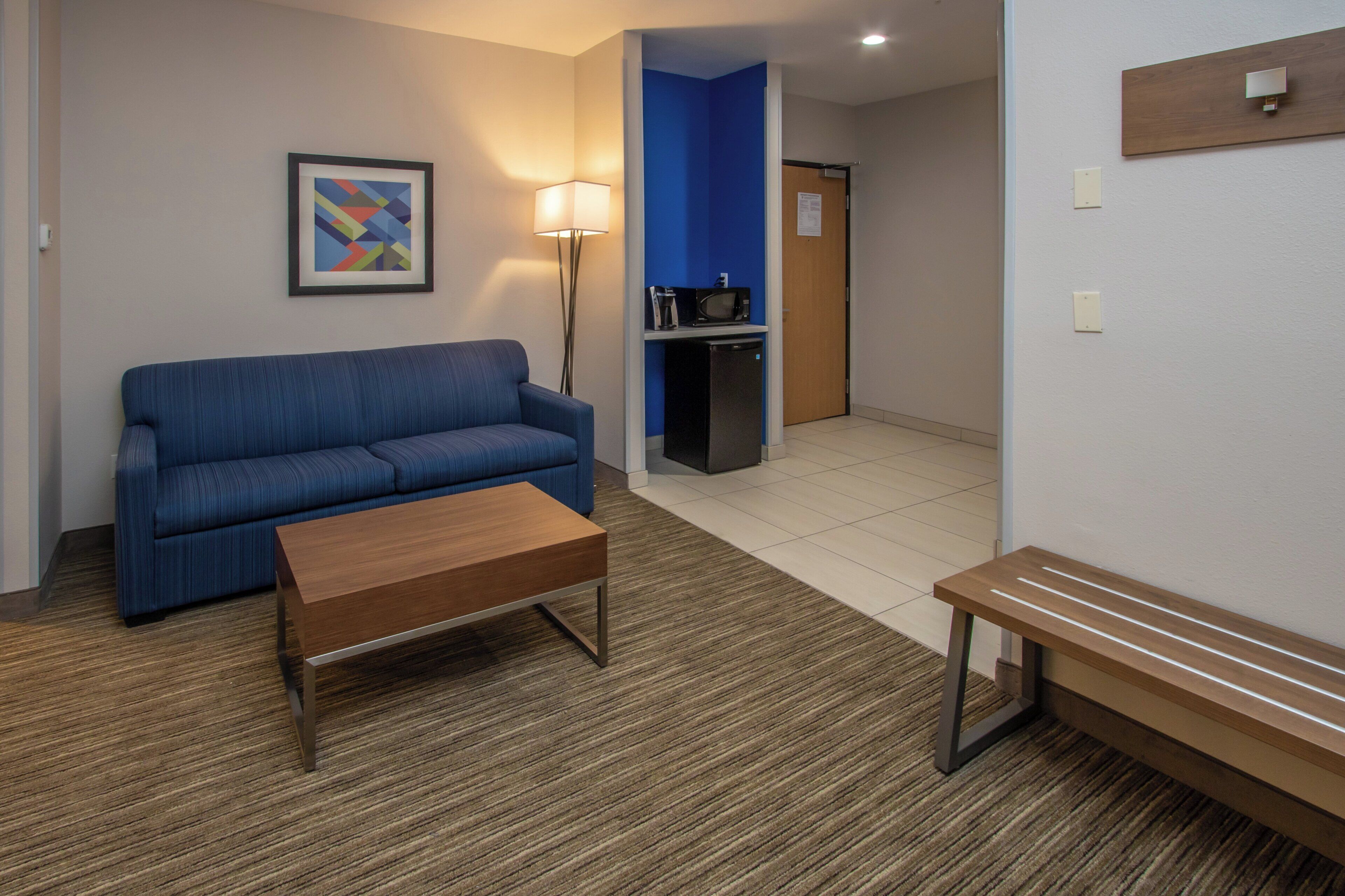 A hotel room with a blue sofa, a wooden coffee table, a bench, a floor lamp, and a kitchenette area.