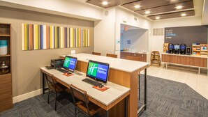 Un espacio de oficina moderno con dos escritorios, cada uno con una computadora portátil y un monitor que muestra 'Holiday Inn Express'. Hay una estación de café con varias cafeteras y un letrero que dice 'NECESIDAD ABSOLUTA: CAFÉ'. La habitación tiene una paleta de colores neutra, una alfombra estampada y un escritorio de madera.