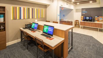Un espace de bureau moderne avec deux bureaux, chacun équipé d’un ordinateur portable et d’un écran affichant le logo « Holiday Inn Express ». On y trouve un coin café avec plusieurs machines à café et un panneau indiquant : « IL FAUT ABSOLUMENT PRENDRE UN CAFÉ ». La pièce est décorée dans des tons neutres, avec une moquette à motifs et un bureau en bois.