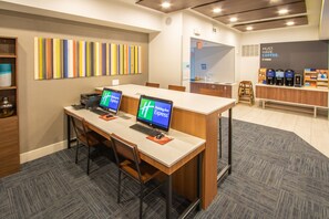 Un espacio de oficina moderno con dos escritorios, cada uno con una computadora portátil y un monitor que muestra 'Holiday Inn Express'. Hay una estación de café con varias cafeteras y un letrero que dice 'NECESIDAD ABSOLUTA: CAFÉ'. La habitación tiene una paleta de colores neutra, una alfombra estampada y un escritorio de madera.