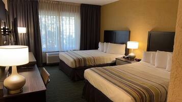 A hotel room with two beds, a desk with a lamp, a window with curtains, and a wall-mounted TV.