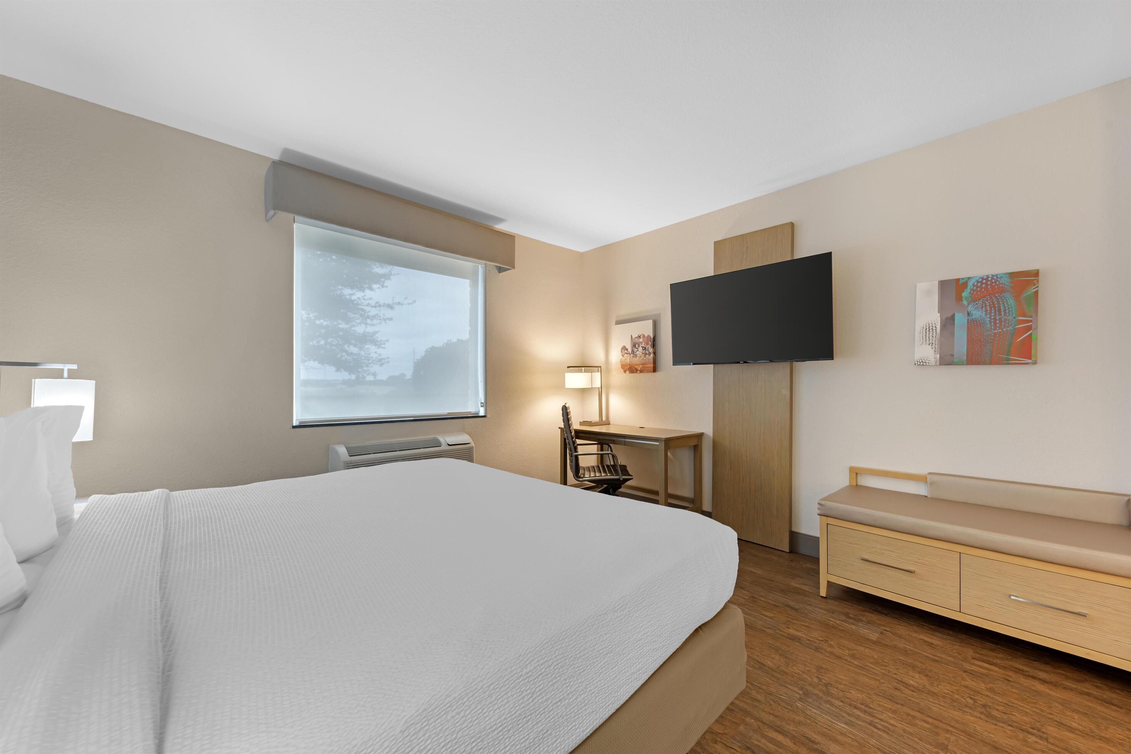 Ett hotellrum med en stor säng, ett skrivbord, en stol, en TV och en bänk.