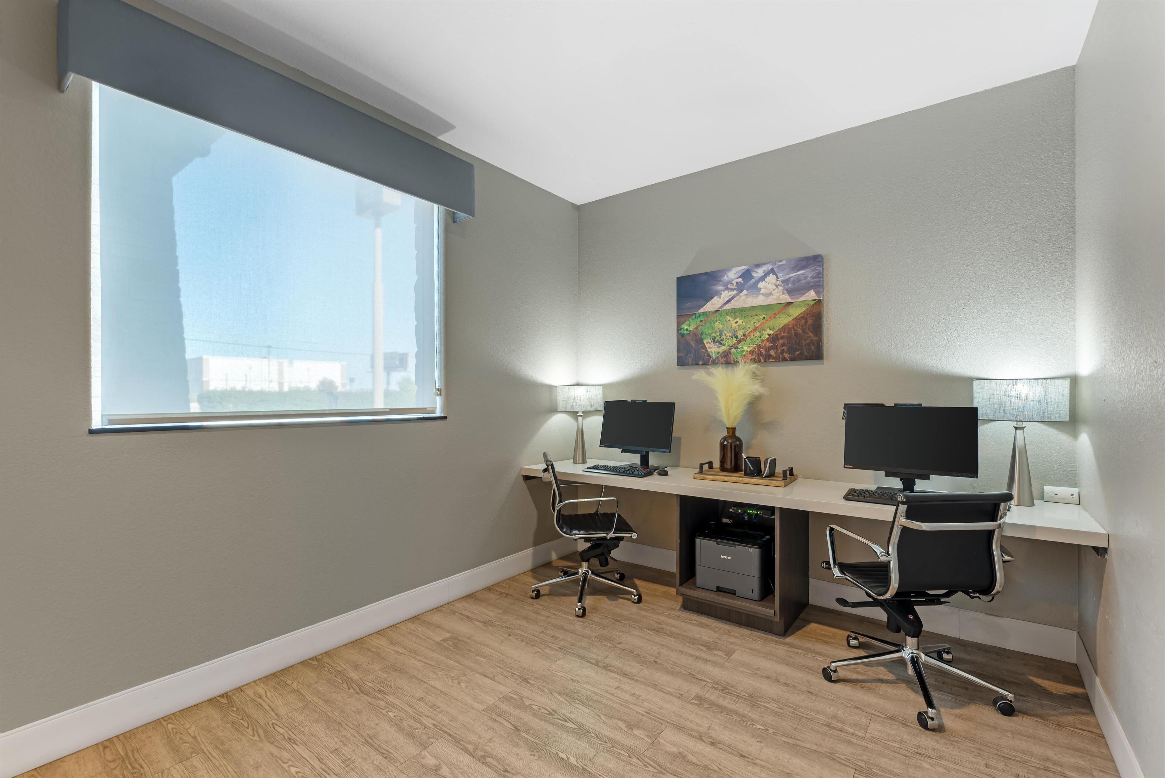 Un espacio de oficina moderno con dos escritorios, cada uno con monitor de ordenador, teclado y silla. Hay una ventana que proporciona luz natural y un cuadro en la pared.