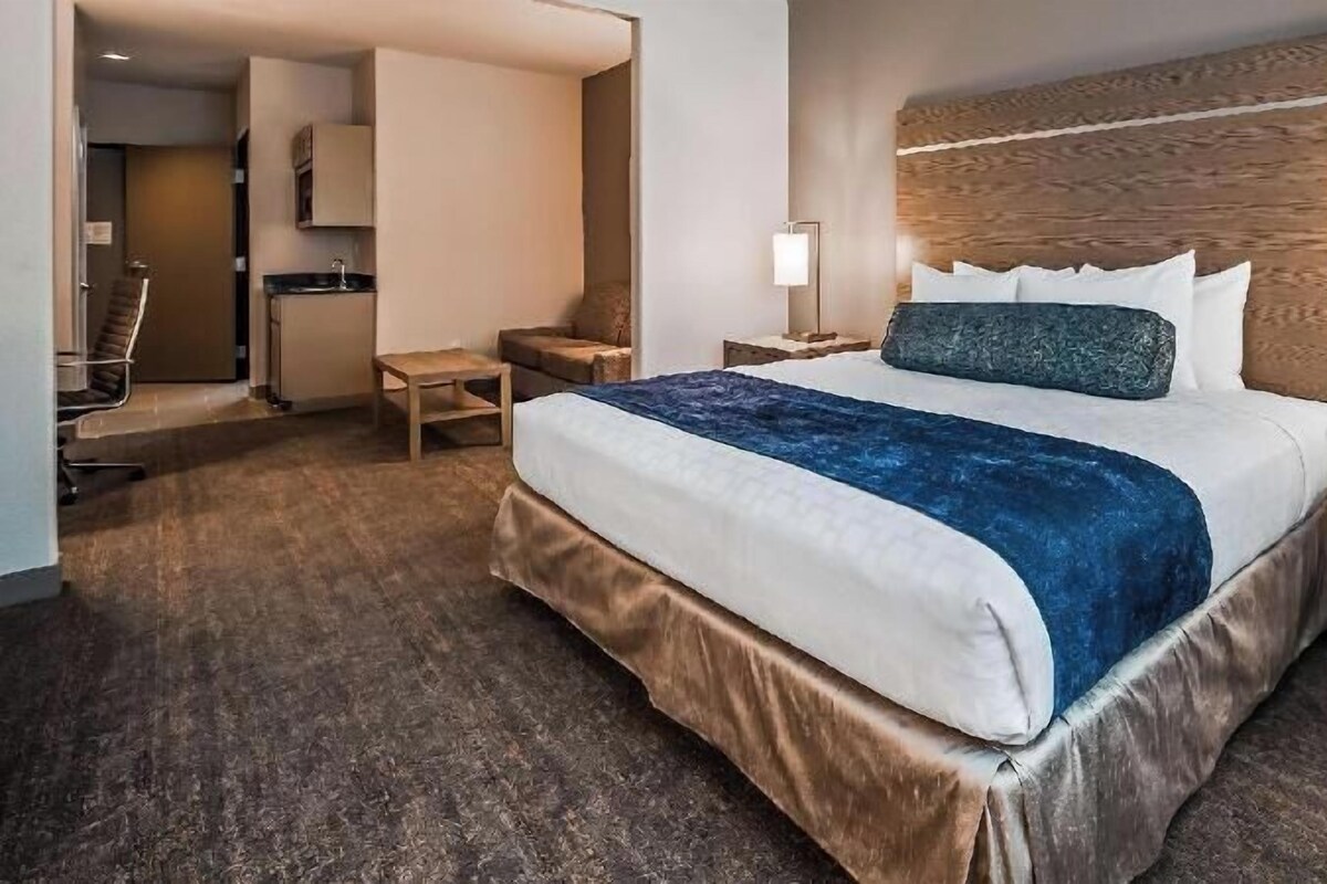 Habitación de hotel con una cama grande, cabecero de madera, un escritorio con lámpara, una silla y una mesita.