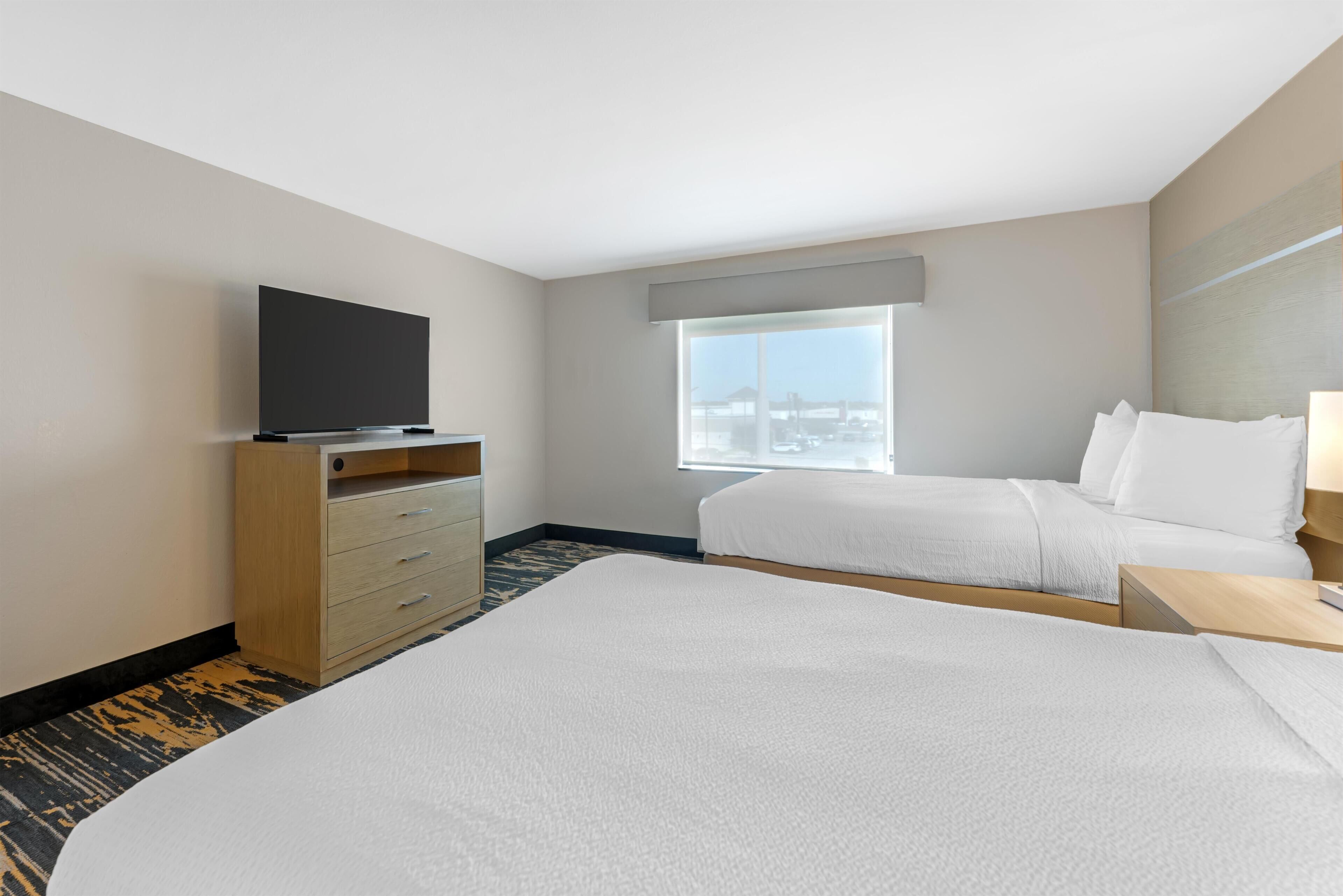 A hotel room with a bed, a TV on a wooden stand, a window with a city view, and a carpeted floor.