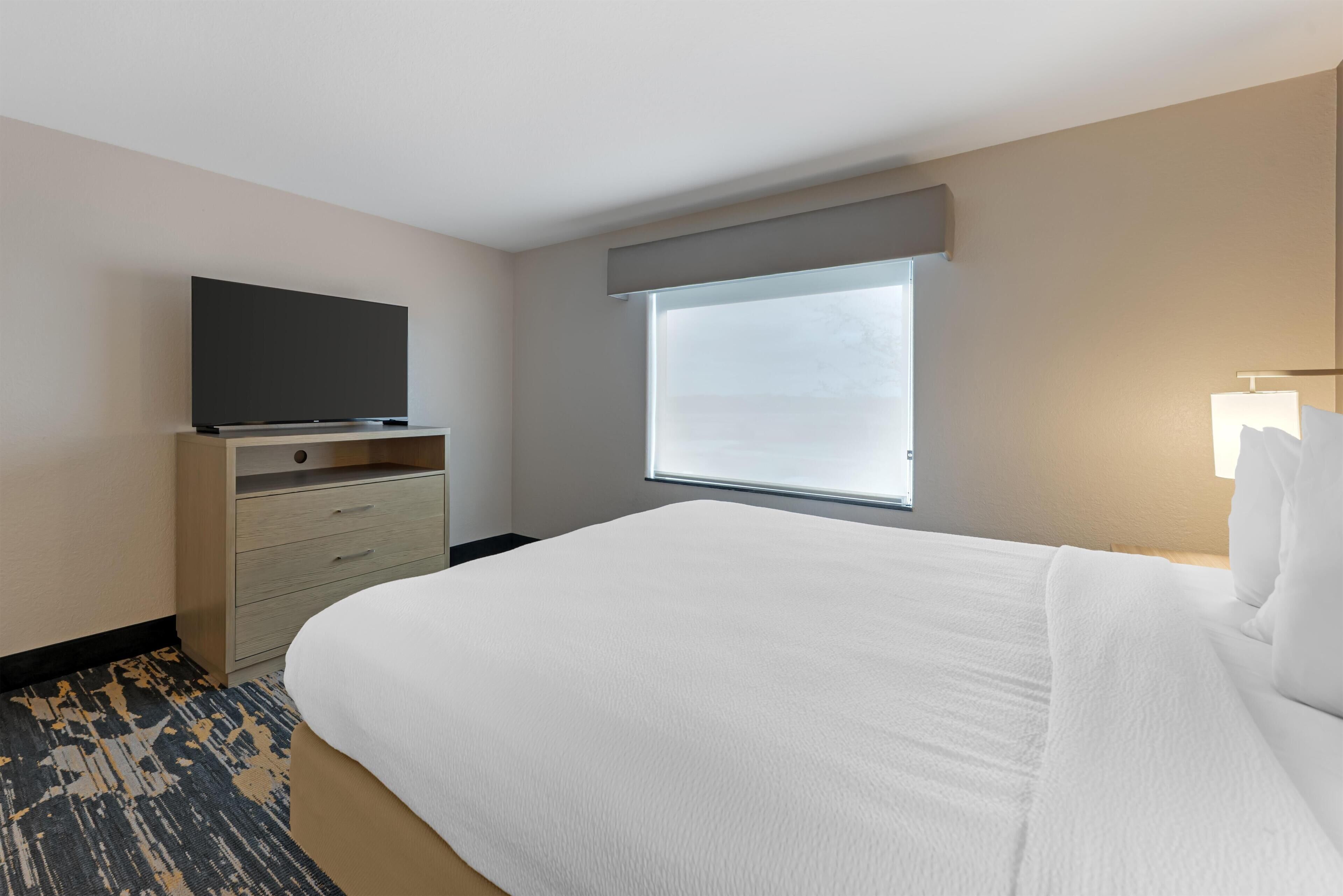 A hotel room with a bed, a flat-screen TV on a wooden dresser, a window with blinds, and a carpeted floor.