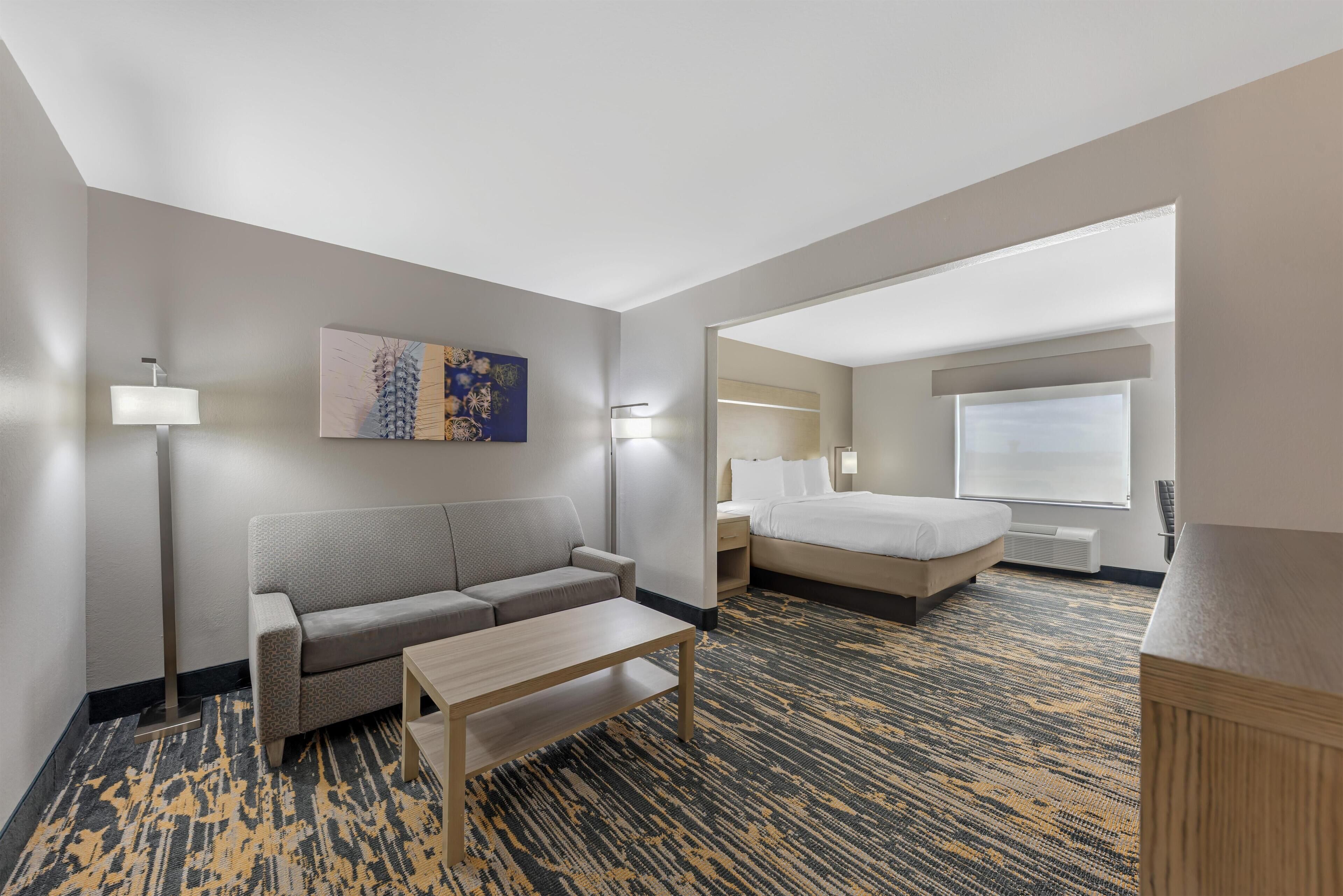 Ein Hotelzimmer mit einer Couch, einem Bett, einem kleinen Tisch, einem Bild an der Wand und einem Fenster mit Jalousien.
