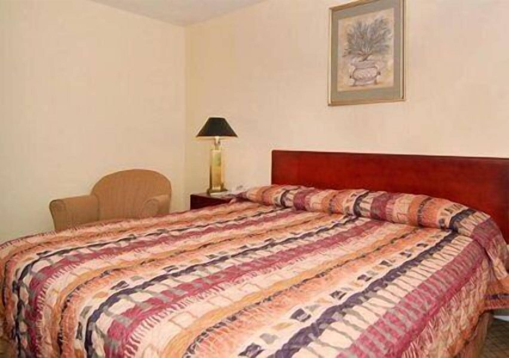 A hotel room with a bed, a chair, a nightstand, a lamp, and a framed picture on the wall.