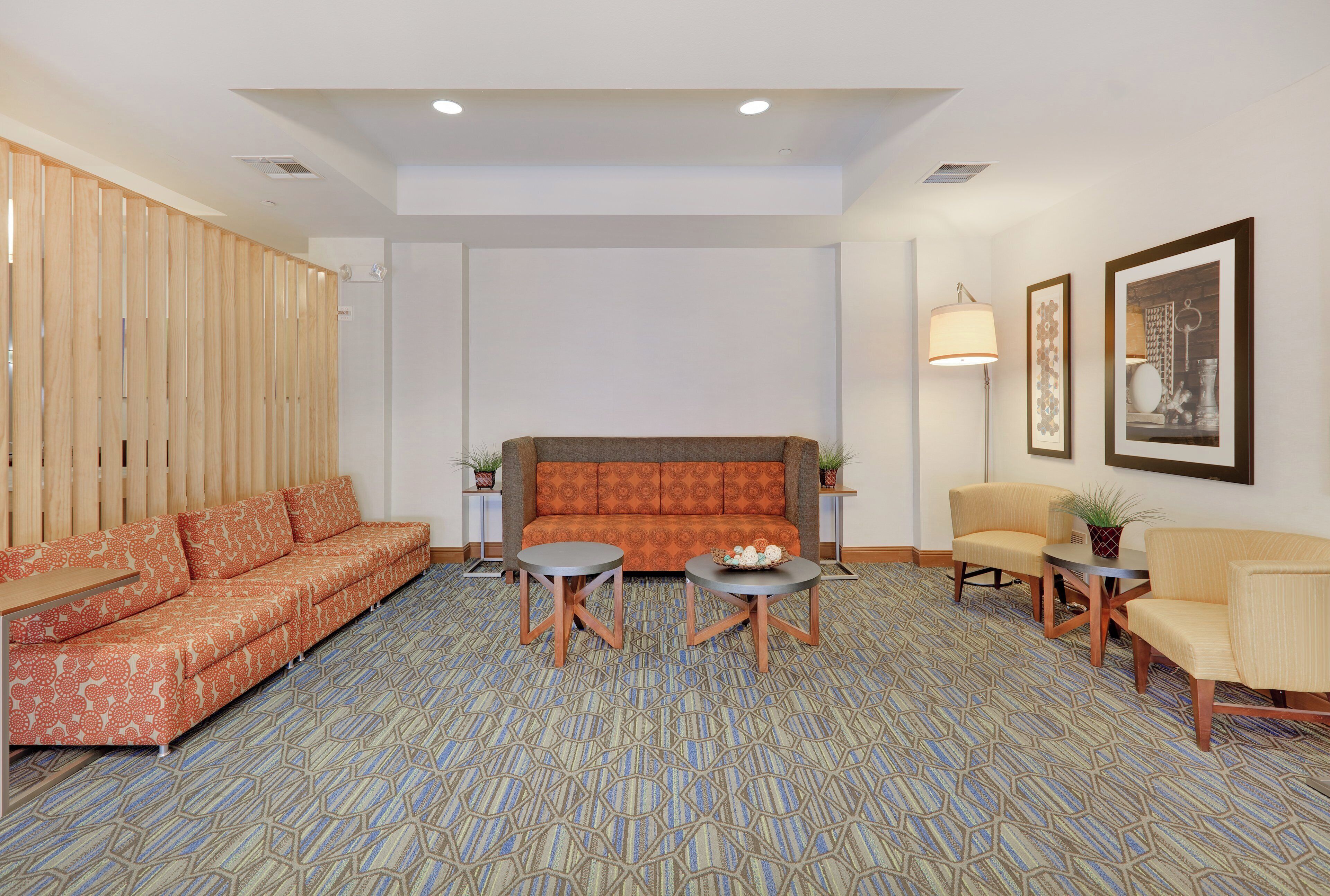 A hotel room with a patterned carpet, a sofa set, two side tables, a floor lamp, and framed artwork on the wall.