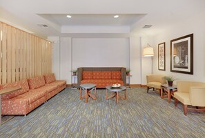Una habitación de hotel con alfombra estampada, un conjunto de sofá, dos mesitas auxiliares, una lámpara de pie y cuadros en la pared.
